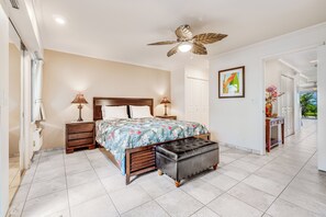 Quarto com cama, mesas de cabeceira, um banco, ventoinha no teto e porta que dá para uma varanda.