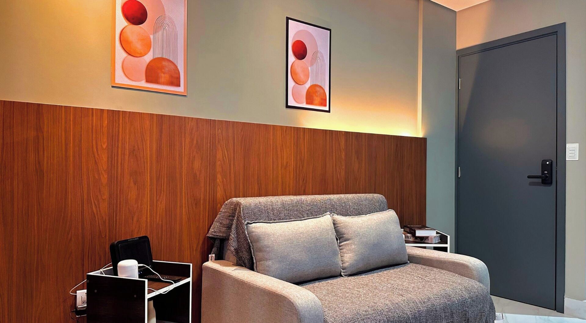 A room with a wooden panel wall, a grey armchair with a pillow, a small table with a laptop, and two framed abstract art pieces on the wall.