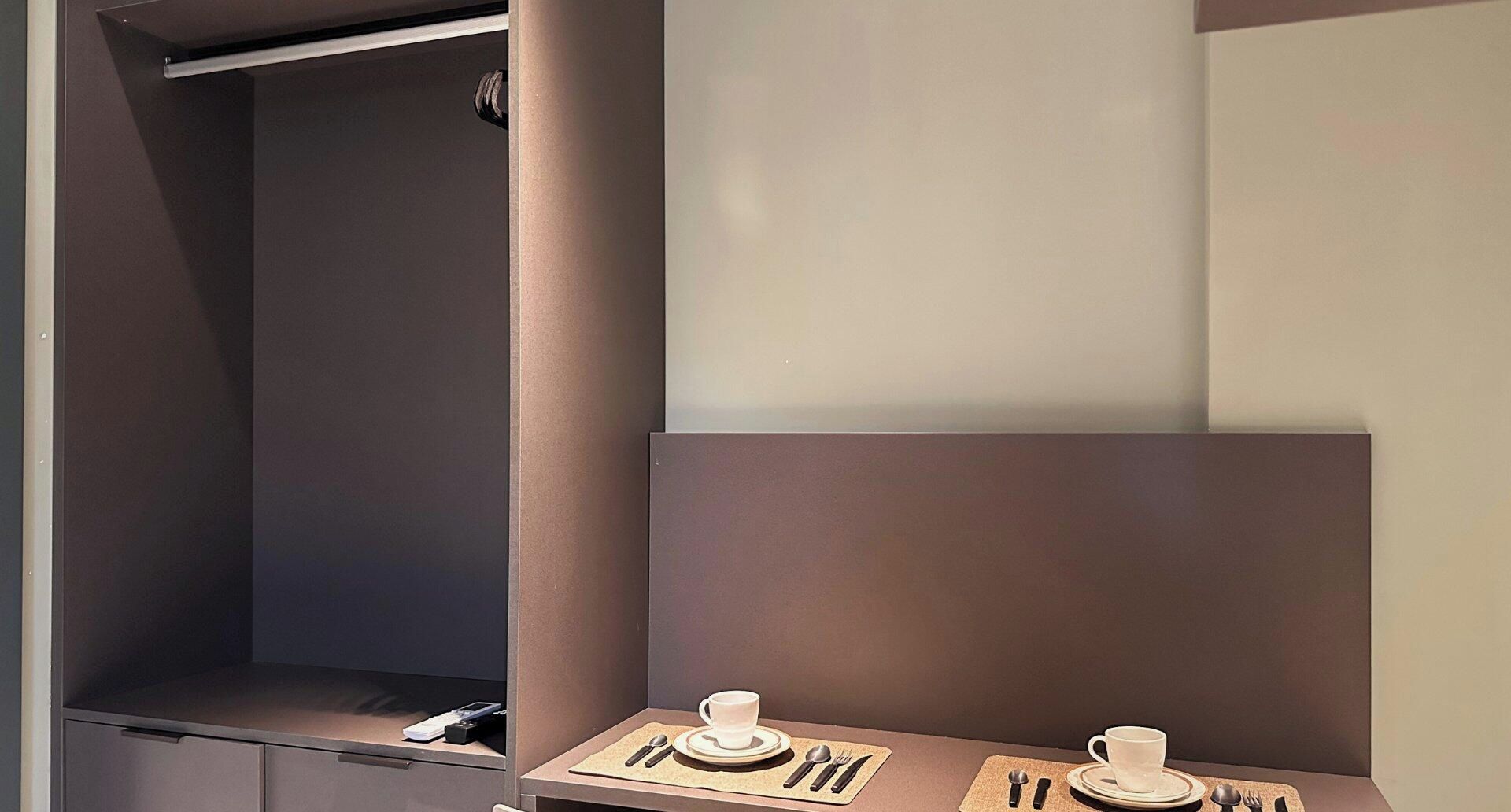 Quarto de hotel com minibar, uma pequena mesa com chávena e talheres, e um guarda-roupa.