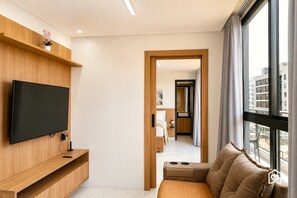 Una habitación de hotel moderna con una televisión de pantalla plana montada en la pared, un estante de madera con una planta en maceta, un sofá con una mesita de centro integrada y un gran ventanal con vistas a la ciudad.