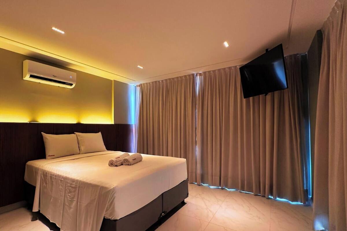 A hotel room with a bed, an air conditioner, a flat-screen TV, and curtains.