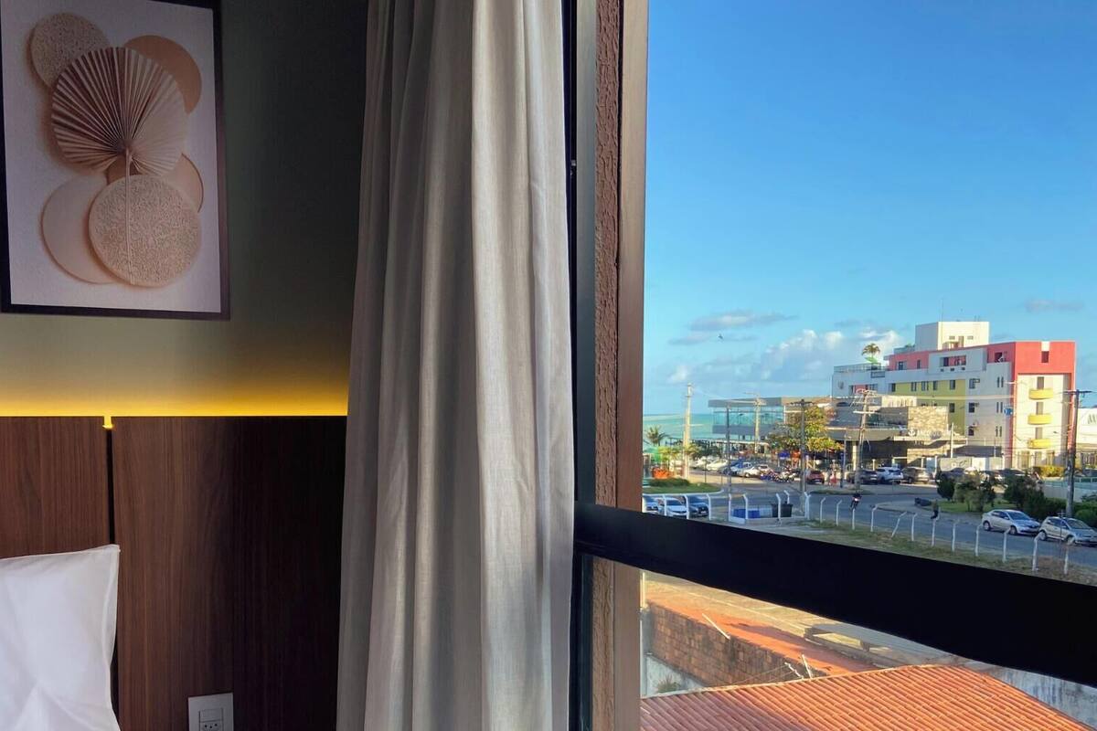 A hotel room with a bed, a window with a view of a cityscape, and a framed artwork on the wall.