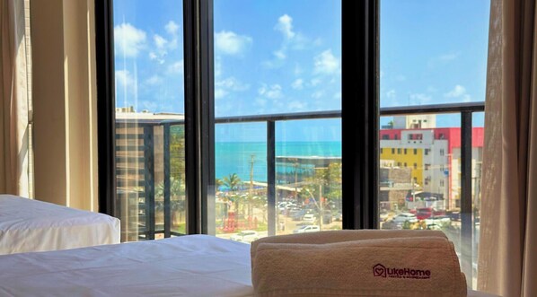 A hotel room with a bed, a window with a view of a beach and cityscape, and a towel with a logo. - Ciel Home - LikeHome Hospedagens (João Pessoa)