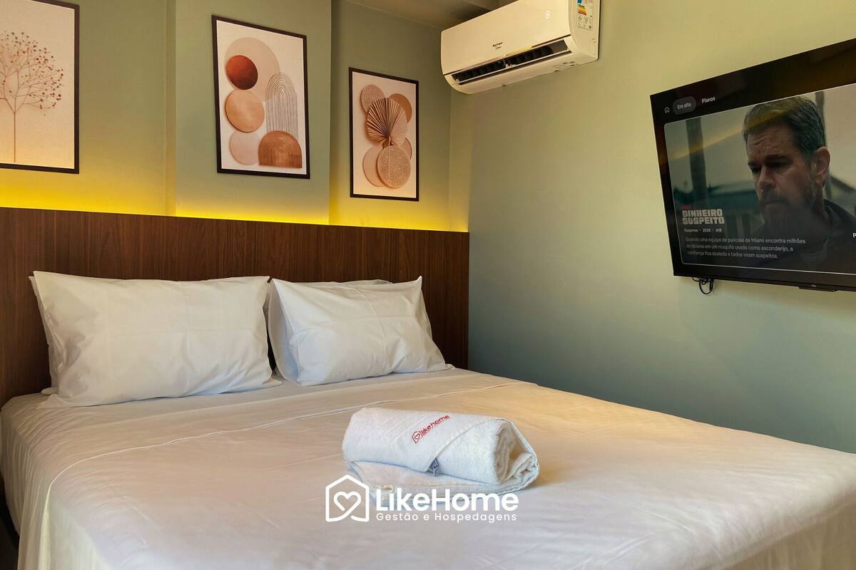 A hotel room with a bed, white pillows, a rolled-up towel, and a TV mounted on the wall displaying a news program.