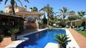 A swimming pool with a house in the background, surrounded by palm trees and a thatched-roof structure. - 4 Bedroom Villa Sleeps 8 With Private Pool (Vera)