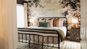 Een slaapkamer met een metalen bedframe, een groot raam met gordijnen, een muur met een boommotief en een nachtkastje met een lamp.