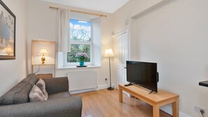 Una sala de estar con un sofá, un televisor sobre un mueble de madera, una ventana con vistas a árboles, y una lámpara sobre una mesita.