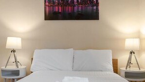 Una cama bien hecha con sábanas y toallas blancas, dos mesitas de noche con lámparas, y un cuadro con una vista de la ciudad en la pared.