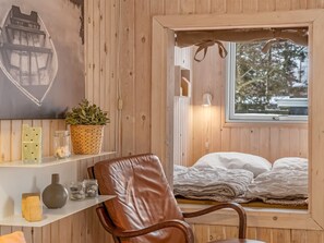 A wooden cabin interior with a bed, a chair, a shelf with decorative items, and a window with a view of trees. - "Irma" - 300m from the sea by Interhome (Hadsund)