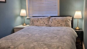 Quarto com colcha de padrão floral, duas luminárias de mesa e uma janela com persianas.