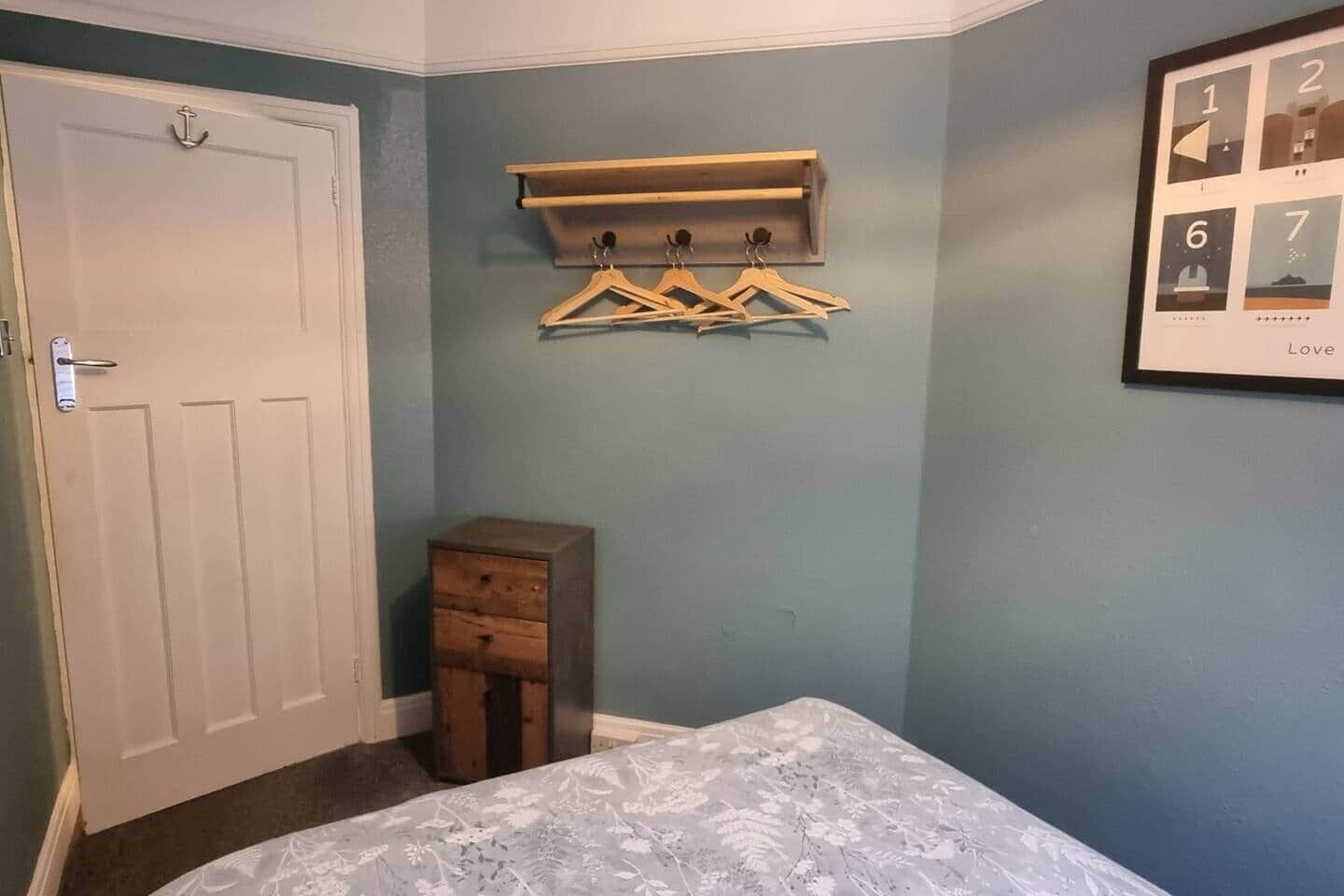 A bedroom with a bed, a wooden dresser, a coat rack with hangers, and a framed picture on the wall.