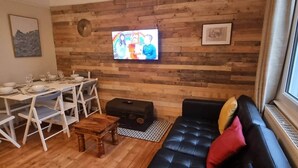 Uma sala de estar com parede de madeira, um sofá de couro preto, um conjunto de mesa de jantar e uma televisão.