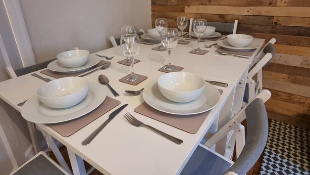 Mesa de jantar posta com pratos brancos, tigelas, talheres e copos de vinho, com parede de madeira e chão ladrilhado.