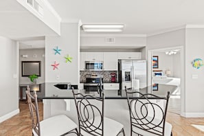 A modern kitchen with white cabinets, a central island, and a dining area with metal chairs. - Splash, Relax & Repeat at Dolphin View Retreat! Beachfront! Sleeps 9! (Panama City Beach)