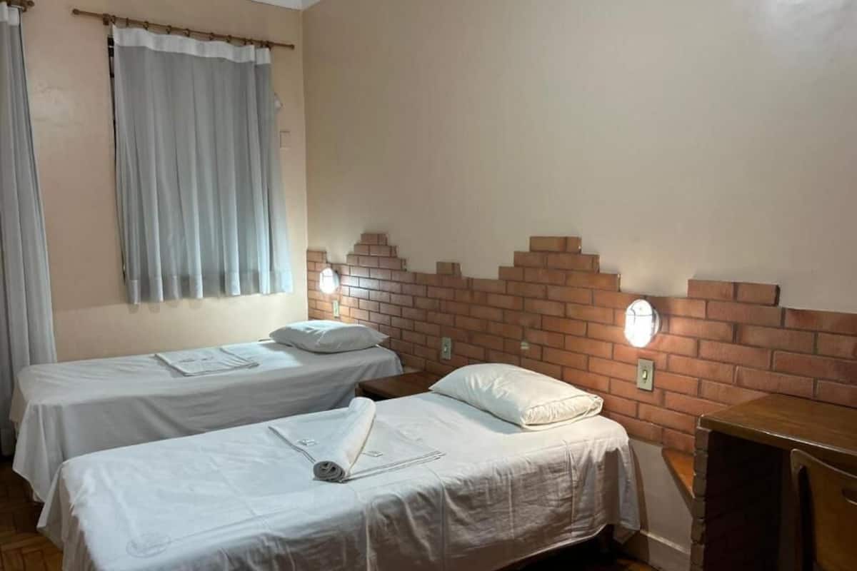 A room with two single beds, a brick accent wall, a small wooden table, and a window with curtains.