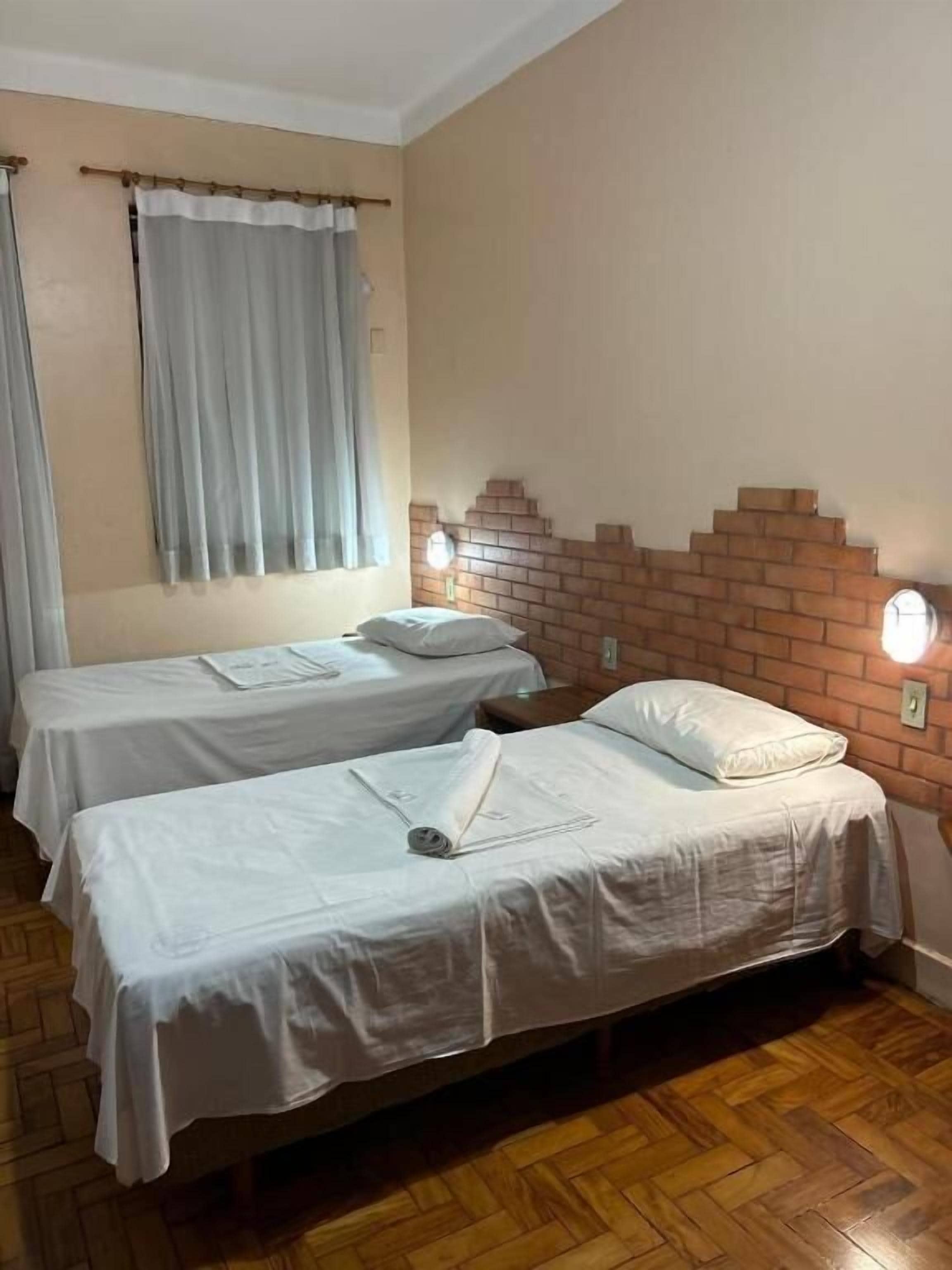Quarto com duas camas de solteiro, parede de destaque em tijolos, janela com cortinas e piso de madeira.