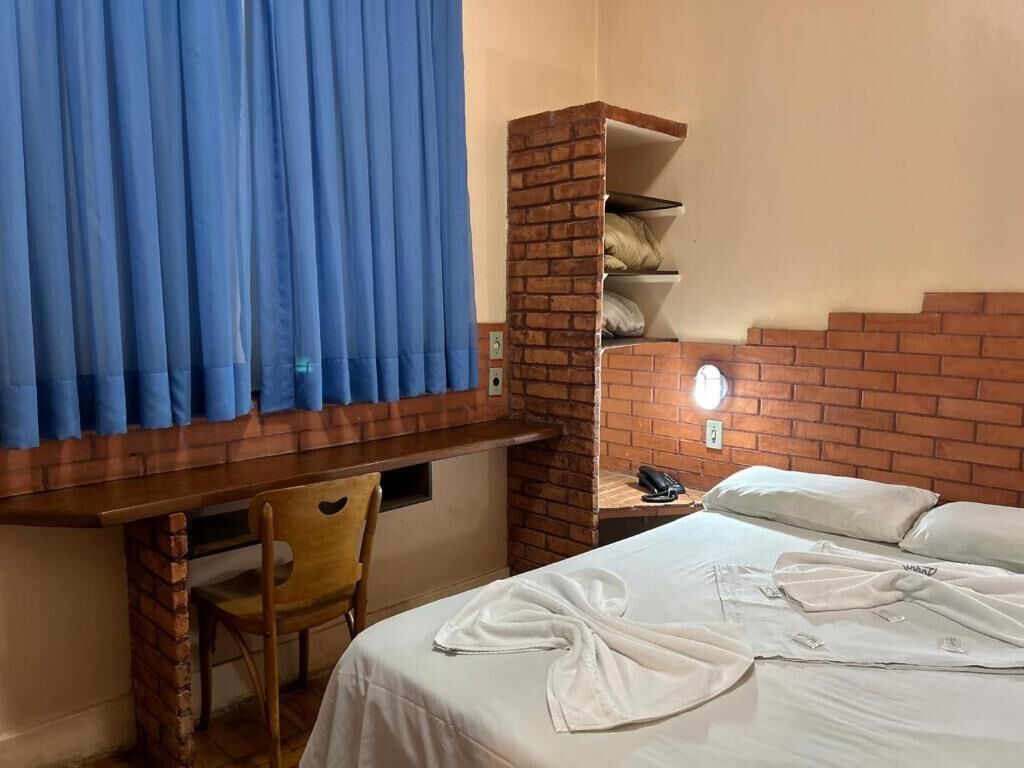 Habitación de hotel con cabecera de ladrillo, escritorio con silla, cama con ropa blanca y cortina azul.