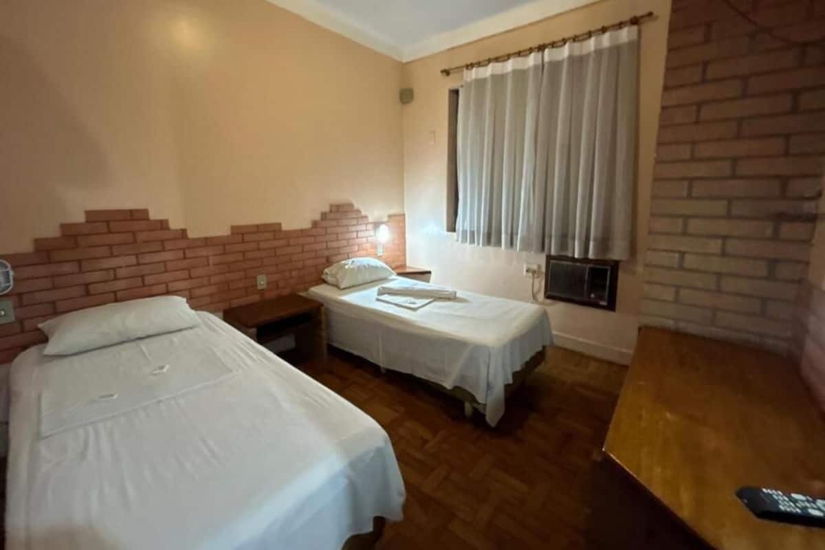 A room with two beds, a brick wall, a window with curtains, and a wooden table.