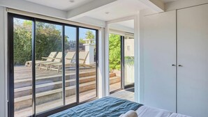 Ein Schlafzimmer mit einem großen Schiebeglasfenster, das auf eine Terrasse mit Liegestühlen führt.