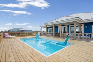 Una terraza de madera con piscina, sillas de descanso y un área cubierta con fachada azul.