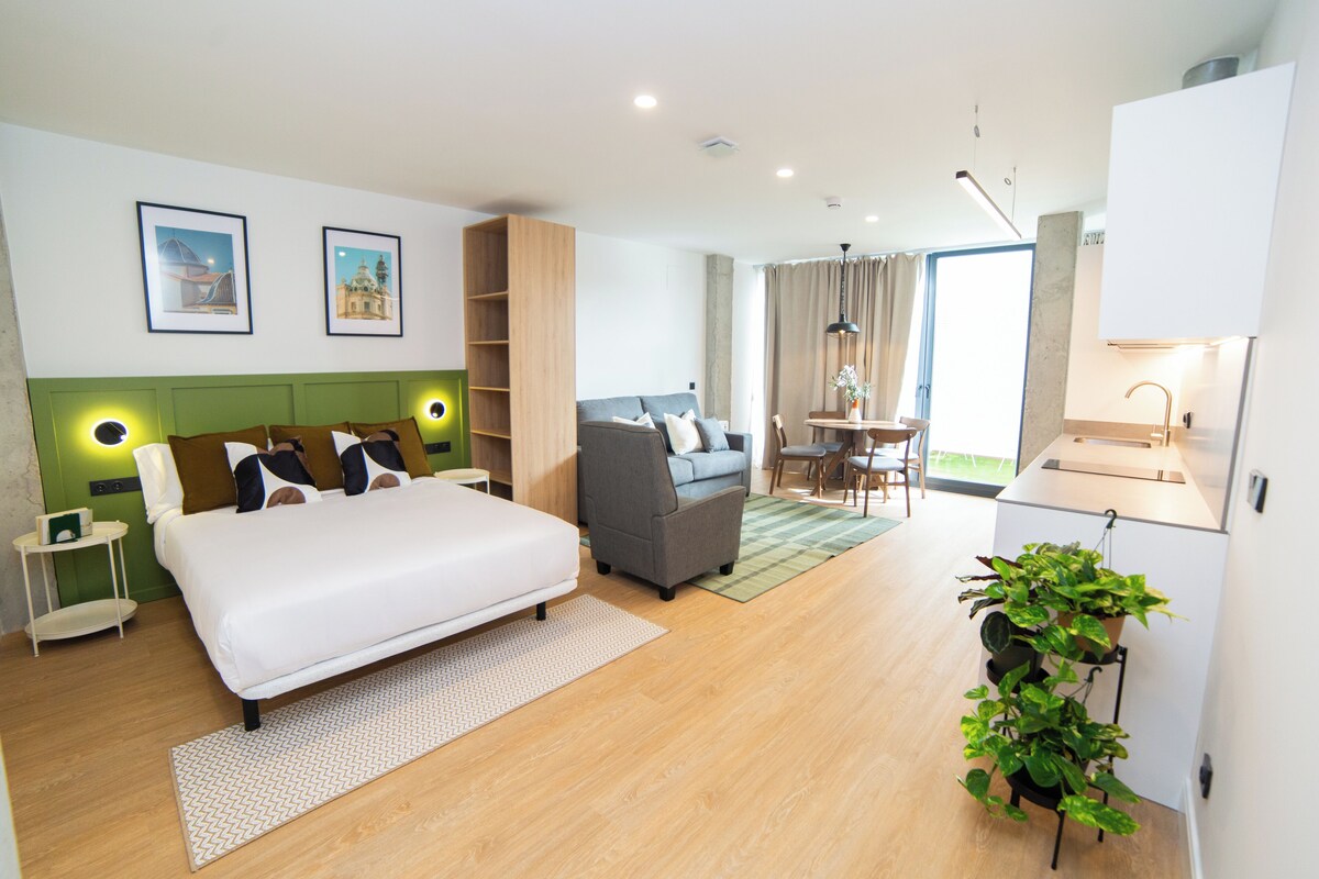 A modern hotel room with a bed, a sofa, a dining area, and a plant.