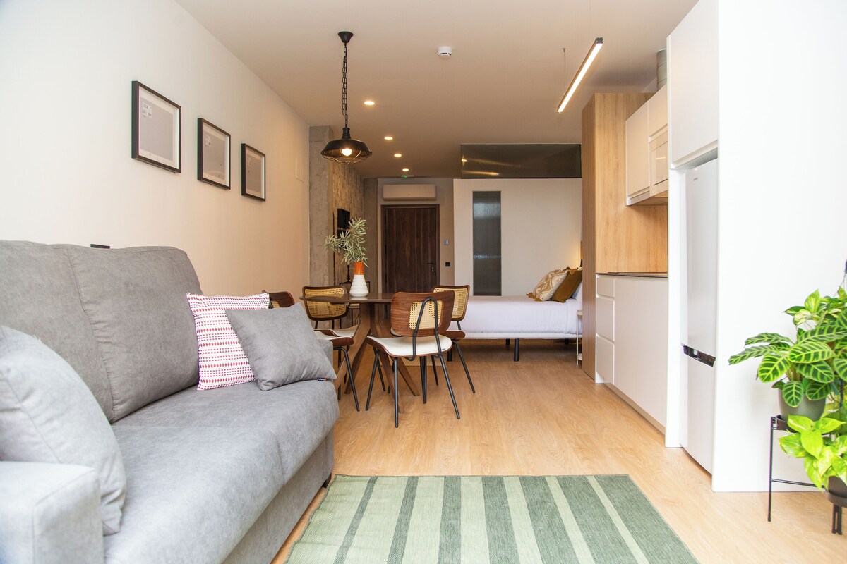 A compact living space with a grey sofa, a dining table with chairs, a small kitchen area, and a bed.