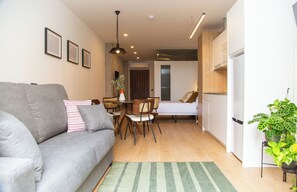 Ein kompakter Wohnbereich mit grauem Sofa, einem Esstisch mit Stühlen, einer kleinen Küchenzeile und einem Bett.