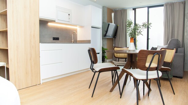 A modern kitchen with white cabinets, a dining area with wooden chairs, a TV, and a large window with curtains. - The 153 Hotel (València)