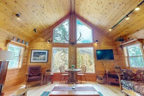 Ein rustikaler Innenraum einer Blockhütte mit Essbereich, einer Couch, einem Fernseher und großen Fenstern.