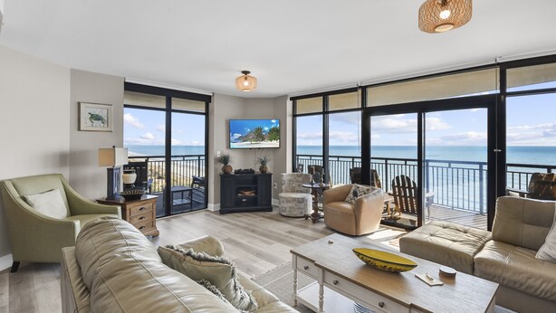 Un salón amplio con un sofá grande, una mesa de centro y un televisor montado en la pared. La habitación tiene ventanas grandes con vista al océano.