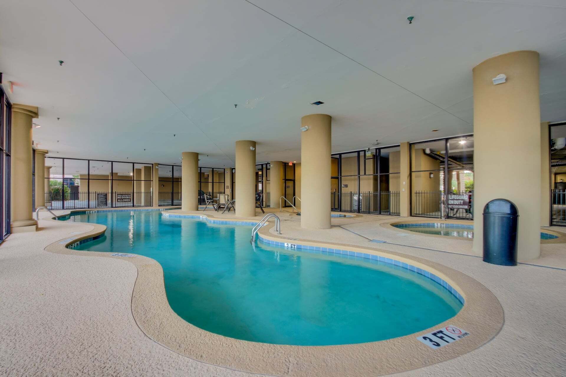 Une piscine intérieure à l’eau claire, entourée de piliers de couleur beige et de murs en verre.