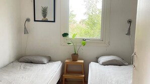 Une petite chambre simple, avec deux lits simples, une fenêtre donnant sur un paysage verdoyant, un tableau représentant un ananas encadré et une plante en pot posée sur un tabouret en bois.
