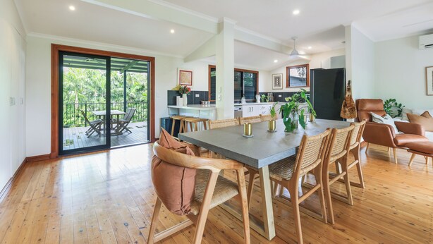 Un vaste espace de salle à manger avec une grande table et des chaises, une cuisine à l’arrière-plan et une porte-fenêtre coulissante donnant sur un balcon.