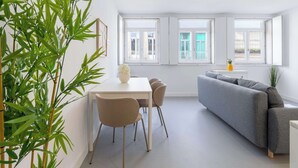 Een moderne woonkamer met een grijze bank, een eettafel met stoelen en een groot raam met uitzicht op een balkon.