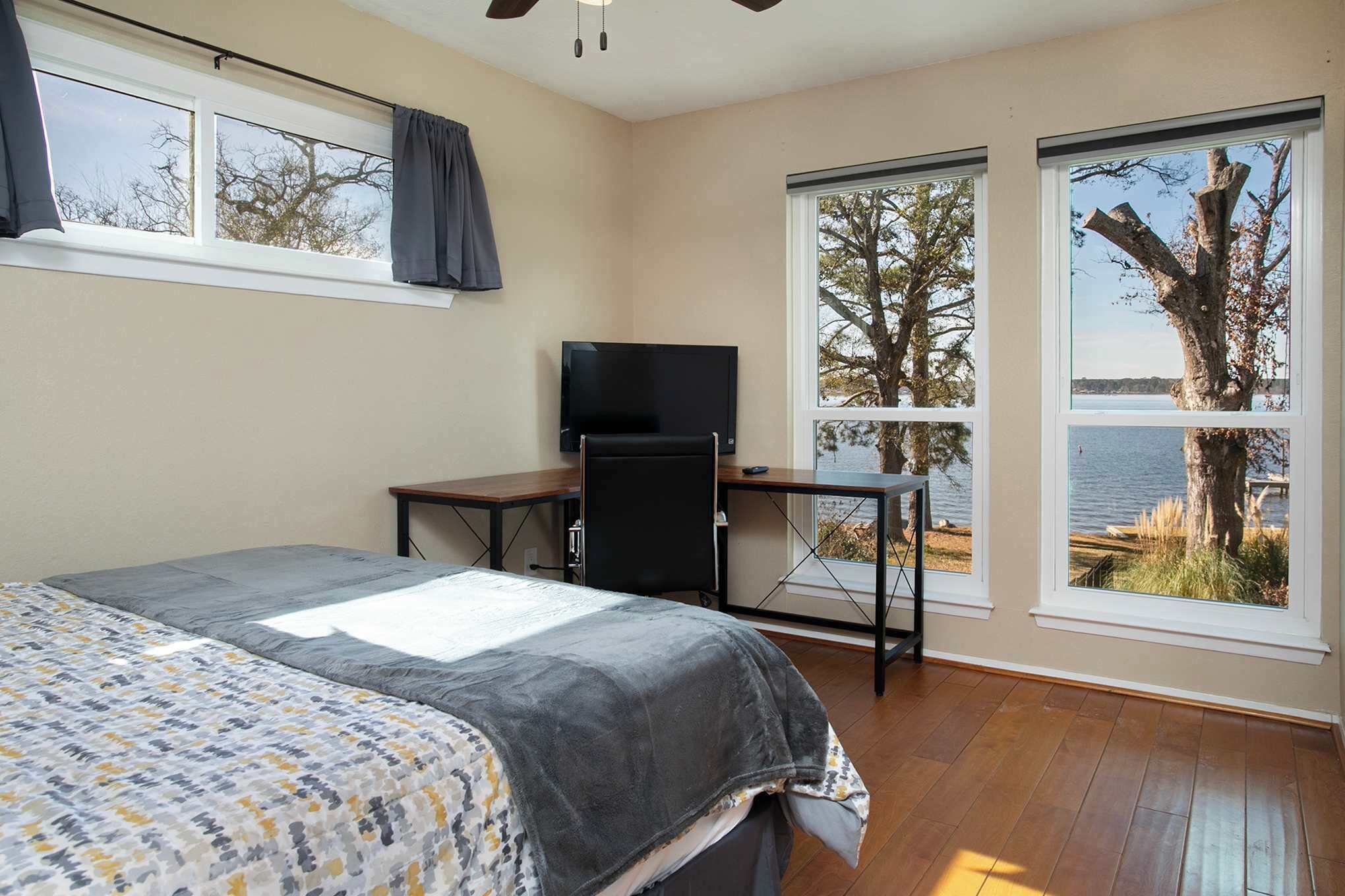 Ein Schlafzimmer mit Bett, Schreibtisch und Fernseher. Große Fenster bieten Blick auf Bäume und Wasser.
