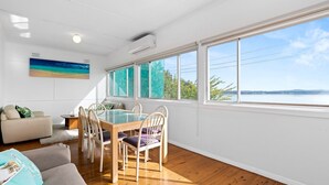 A bright room with a wooden floor, a dining table with chairs, a sofa, and a large window with a view of the water and trees. - Lakeside Cottage Escape - Peaceful Stay with Scenic Lake Views (Toukley)