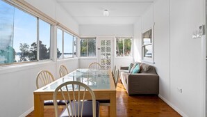 A dining area with a glass table, wooden chairs, and a sofa with cushions. - Lakeside Cottage Escape - Peaceful Stay with Scenic Lake Views (Toukley)