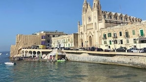 Un edificio costero con una torre destacada y arcos, junto a una playa con gente y un bote verde.