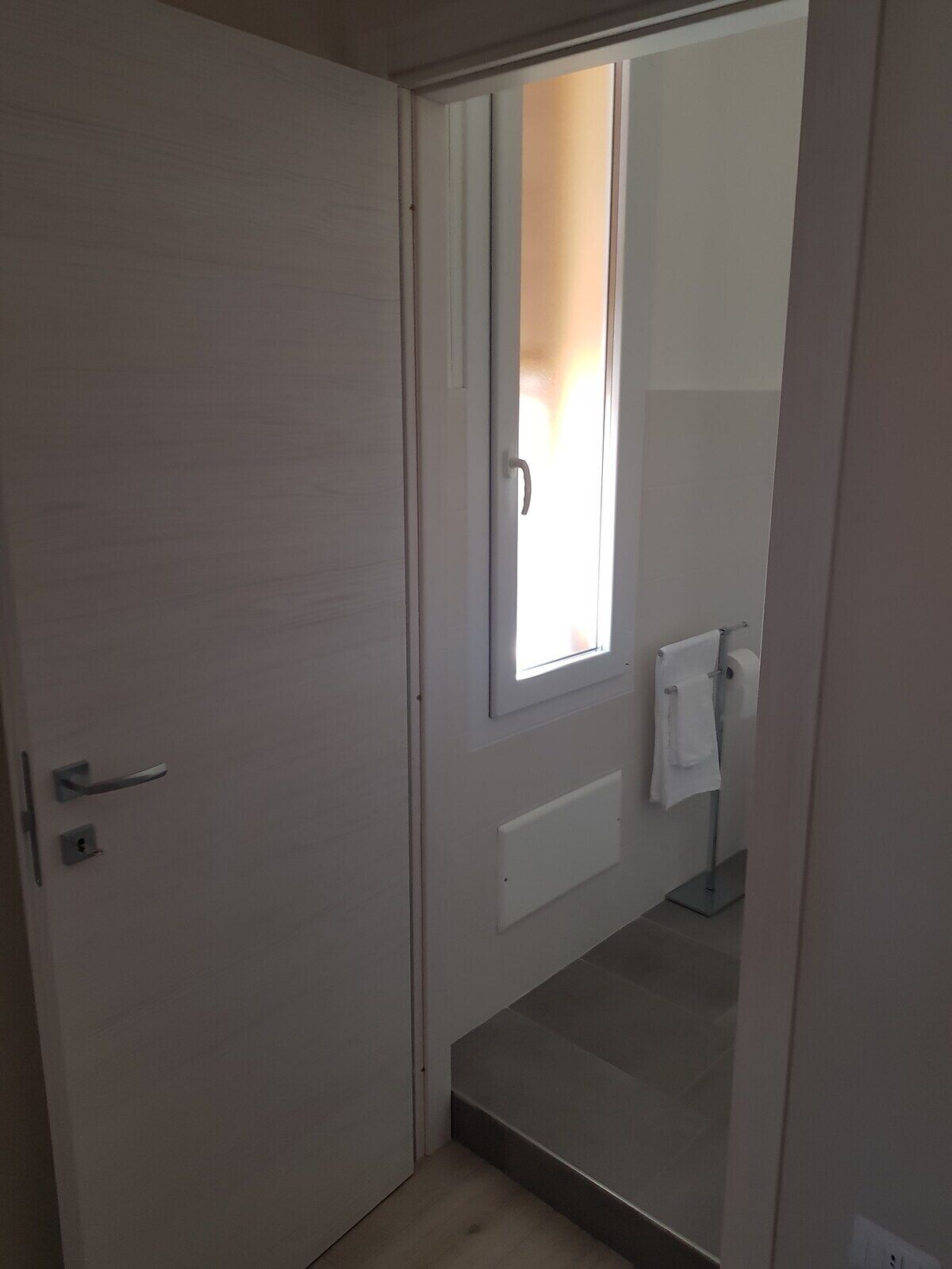 A room with a door leading to a bathroom. The bathroom has a toilet, a towel rack, and a window.