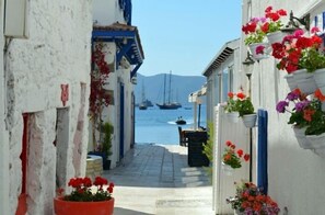 A narrow street with white buildings, potted flowers, and boats in the background. - Paradise Garden Bodrum (Bodrum)