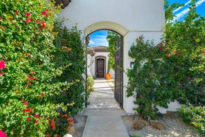 A pathway leading to a building with a brown door, surrounded by lush greenery and flowering plants. - Stunning Private Pool Home Overlooking the 8th and 9th Double Fairway in PGA (La Quinta)