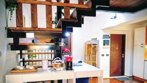 Cozinha com bancada branca, prateleiras de madeira e uma planta pendente. Há uma cadeira azul e um armário de madeira ao fundo.