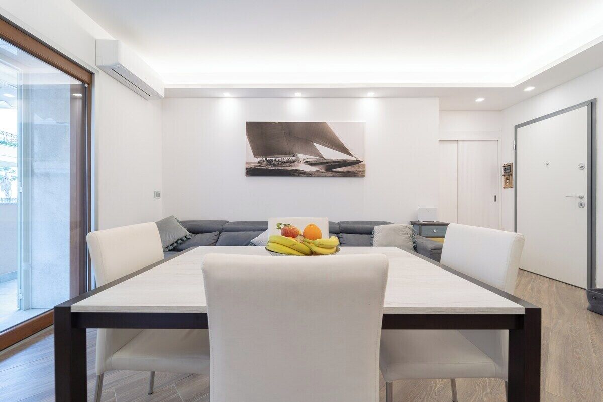 A modern dining area with a large table, white chairs, a gray sofa, and a wall-mounted artwork of a sailboat.