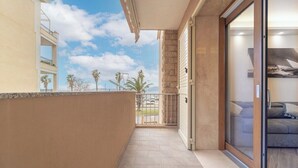 Un balcón con vistas a palmeras y montañas, una puerta corredera de vidrio que da a una habitación con un sofá y un suelo de baldosas.