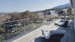 A wooden deck with outdoor furniture, a glass railing, and a view of a coastal town and mountains. - Villa with sea and mountain views (FURIANI)
