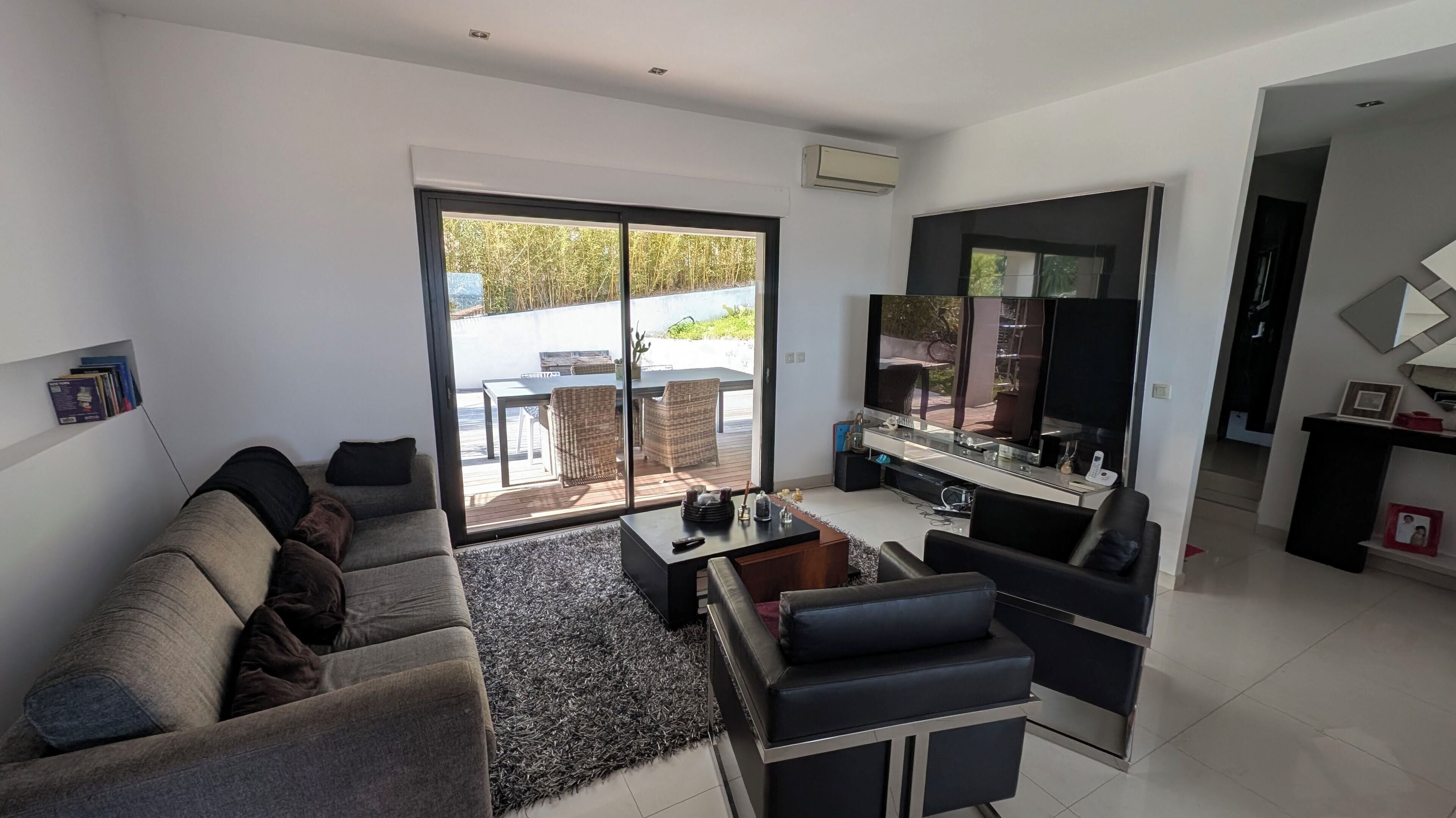 Una sala de estar moderna con un sofá gris, una mesa de centro y un televisor. Hay una puerta corrediza de vidrio que da a un área exterior con una mesa y sillas.