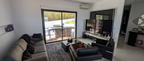 Una sala de estar moderna con un sofá gris, una mesa de centro y un televisor. Hay una puerta corrediza de vidrio que da a un área exterior con una mesa y sillas.