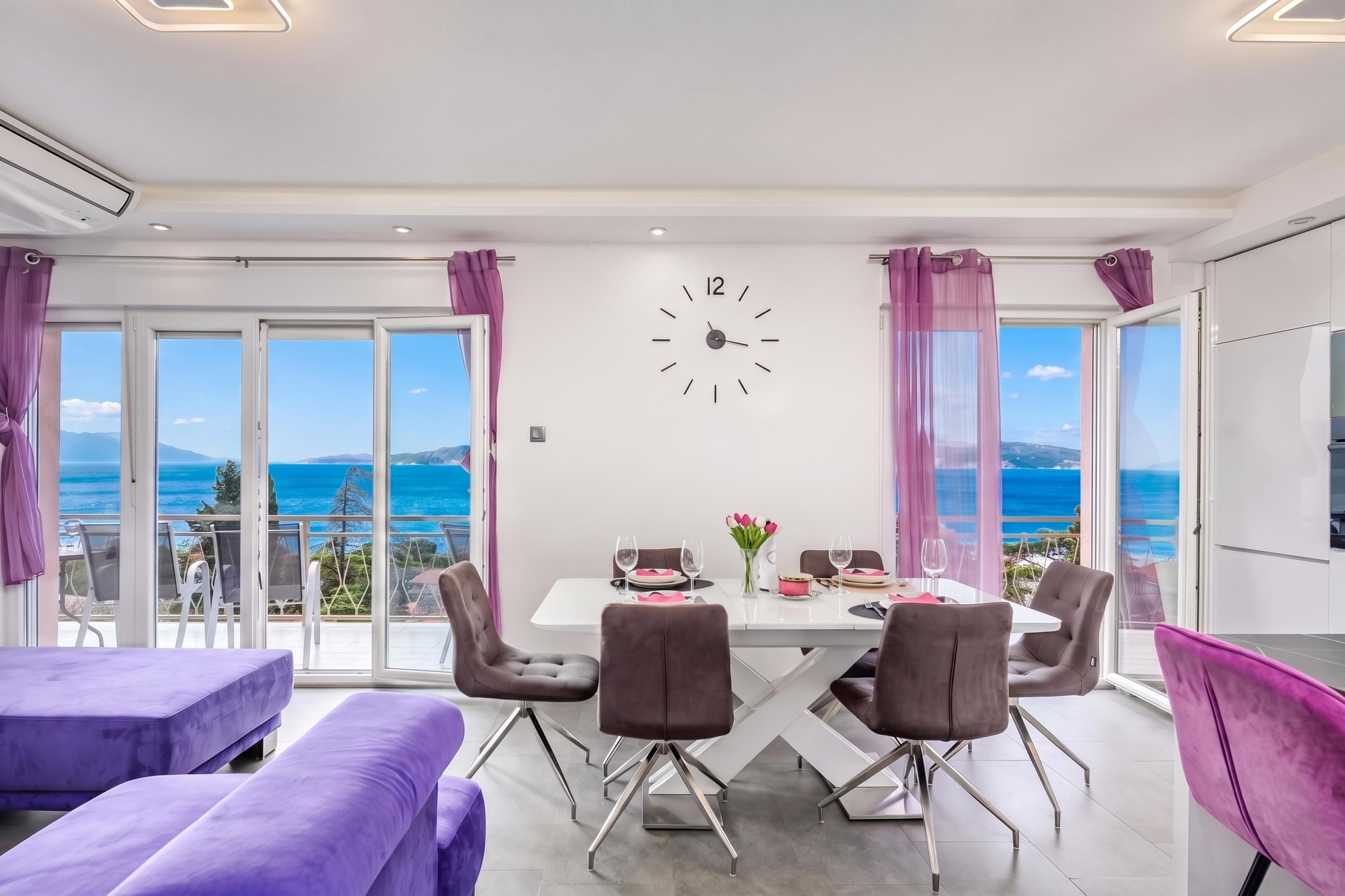 A dining area with a table set for four, a clock on the wall, and a view of the sea.