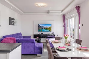 Una sala de estar moderna con un sofá morado, una mesa de comedor para dos personas y un televisor de pantalla plana montado en la pared.