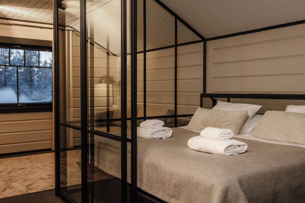A modern hotel room with a glass partition, a bed with neatly folded towels, and a view of a snowy landscape through a window.
